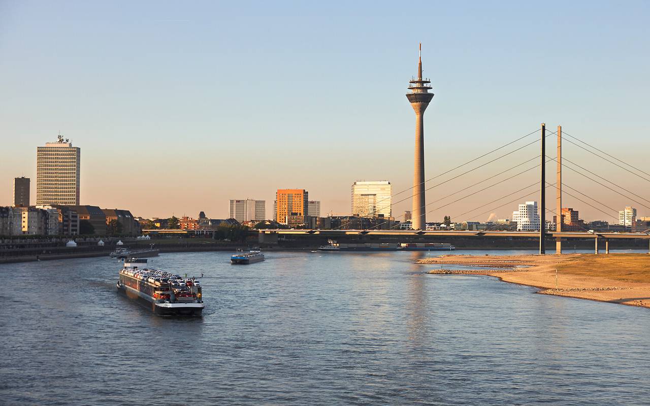 Links und im Vordergrund der Rhein, in dem ein Containerschiff schwimmt, rechts das Ufer. Im Hintergrund der Blick auf Düsseldorf, die Rheinkniebrücke und den Rheinturm.