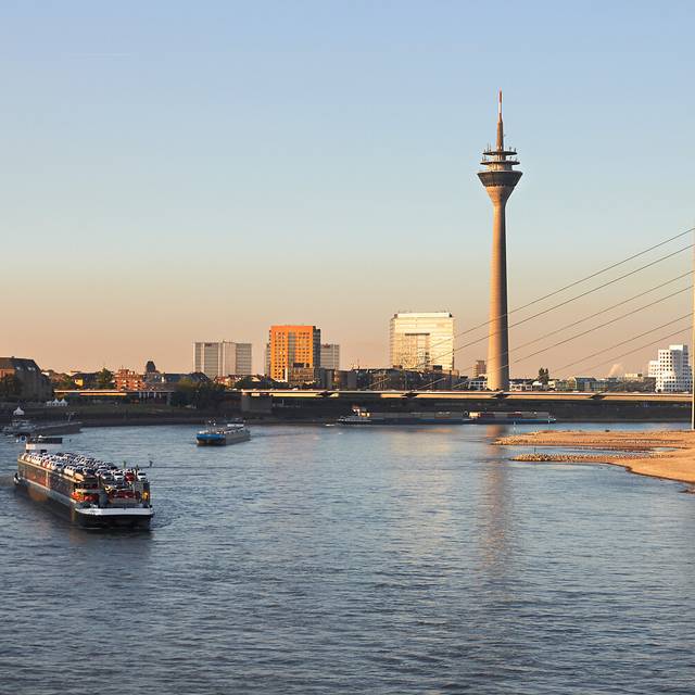 Links und im Vordergrund der Rhein, in dem ein Containerschiff schwimmt, rechts das Ufer. Im Hintergrund der Blick auf Düsseldorf, die Rheinkniebrücke und den Rheinturm.