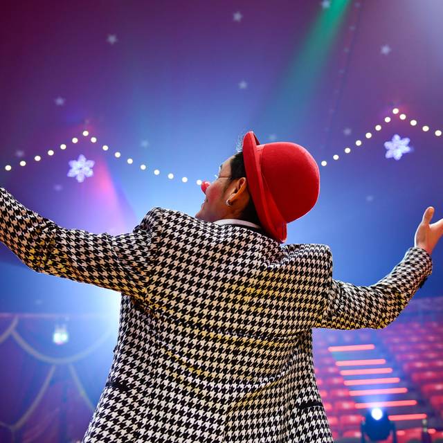 Ein Clown steht in der Manege und breitet die Arme aus, man sieht ihn von hinten