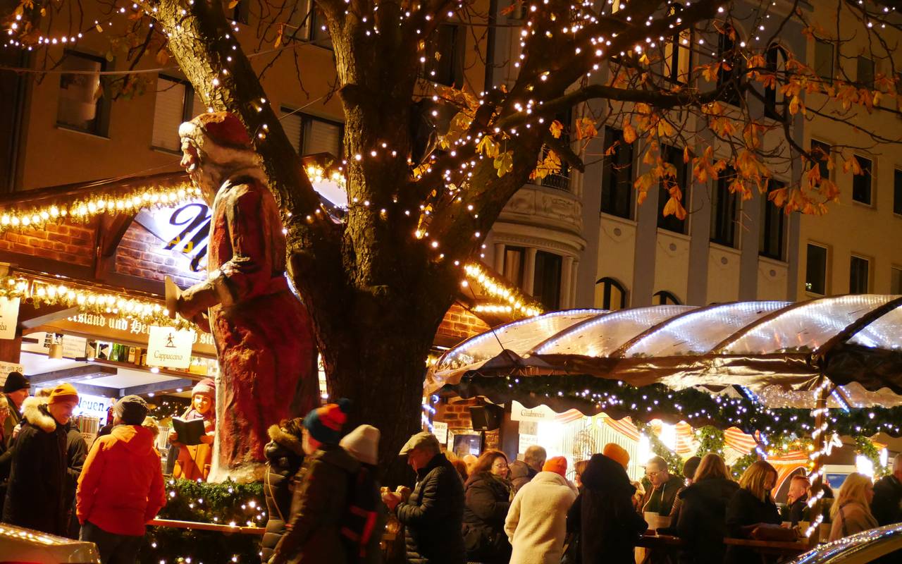 Menschen stehen in Winterkleidung im halbdunkel vor beleuchteten Buden. In der Mitte ein weihnachtlich geschmückter Baum.