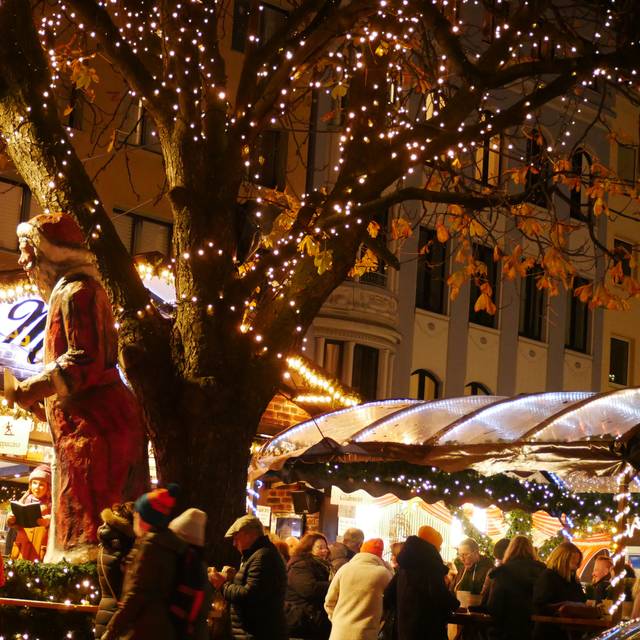 Menschen stehen in Winterkleidung im halbdunkel vor beleuchteten Buden. In der Mitte ein weihnachtlich geschmückter Baum.