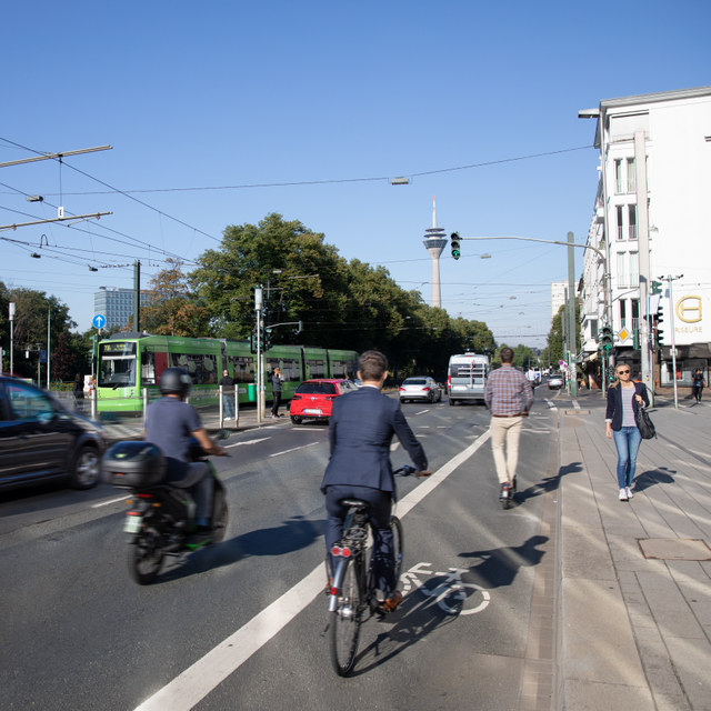 Das Foto zeigt verschiedene Formen der Mobilität am Graf-Adolf-Platz. Neben der Bahn fahren Autos. Außerdem sind Radfahrer auf einem Fahrradstreifen unterwegs und Fußgänger laufen über den Bürgersteig.