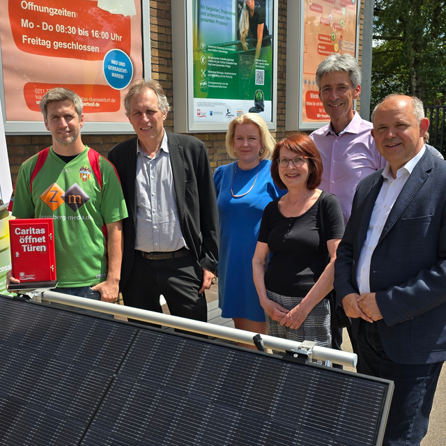 Sechs Menschen der Stadt, Caritas, Bürgerstiftung und Deutschen Postcode Lotterie stehen hinter einem Solarpanel. Einer von ihnen hält einen Flyer hoch.