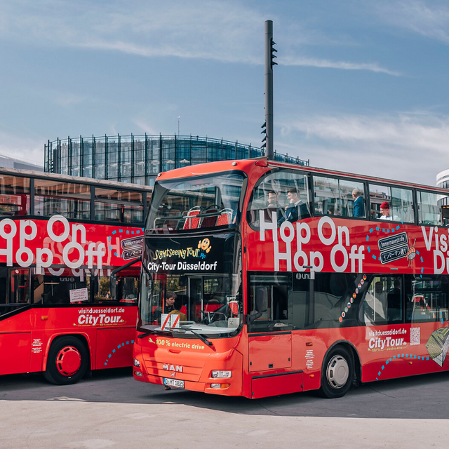 Blick auf zwei elektrische Hop-on-hop-off-Busse. Die Busse sind rote Doppeldecker-Busse mit Düsseldorf-Symbolen.