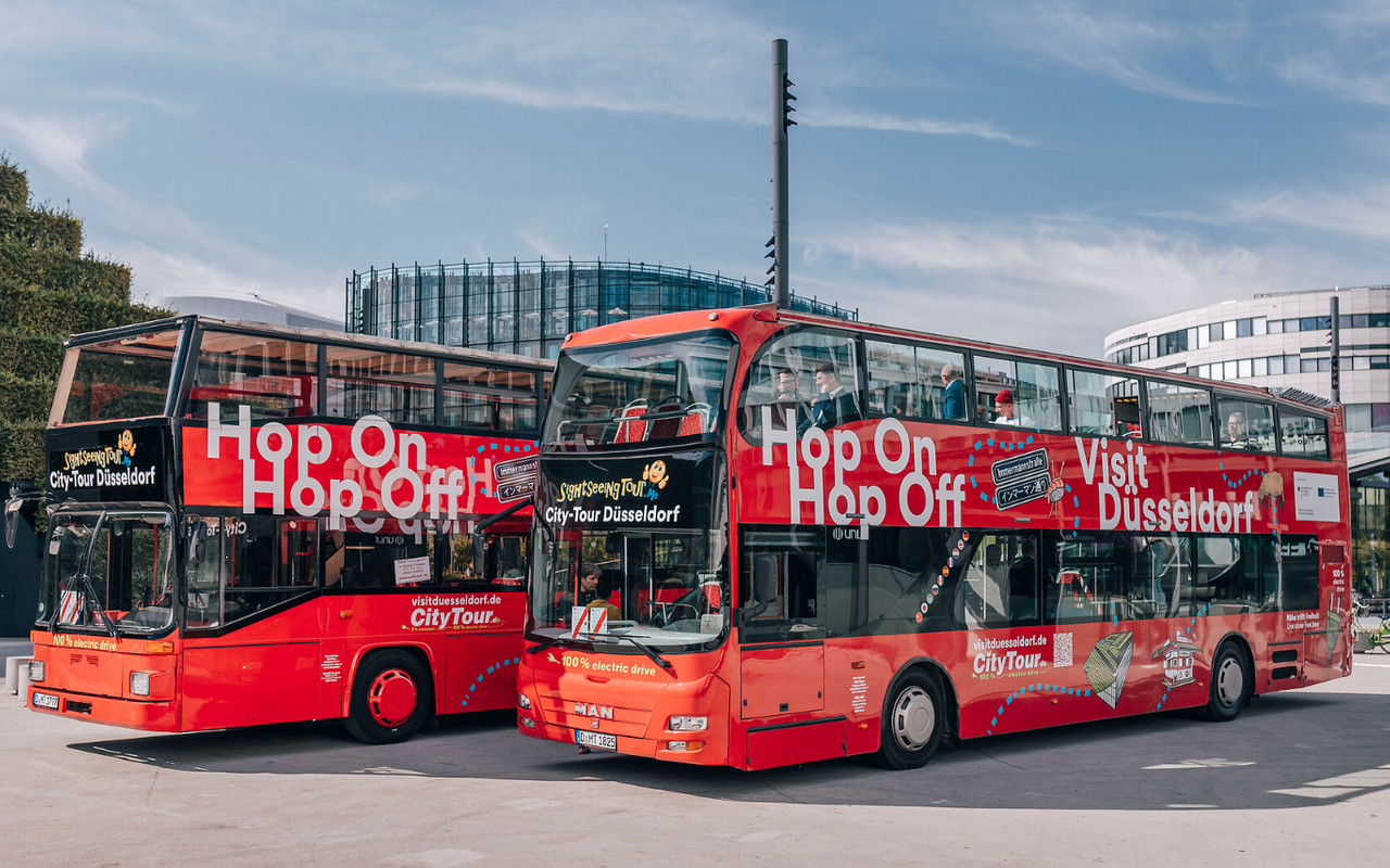 Blick auf zwei elektrische Hop-on-hop-off-Busse. Die Busse sind rote Doppeldecker-Busse mit Düsseldorf-Symbolen.
