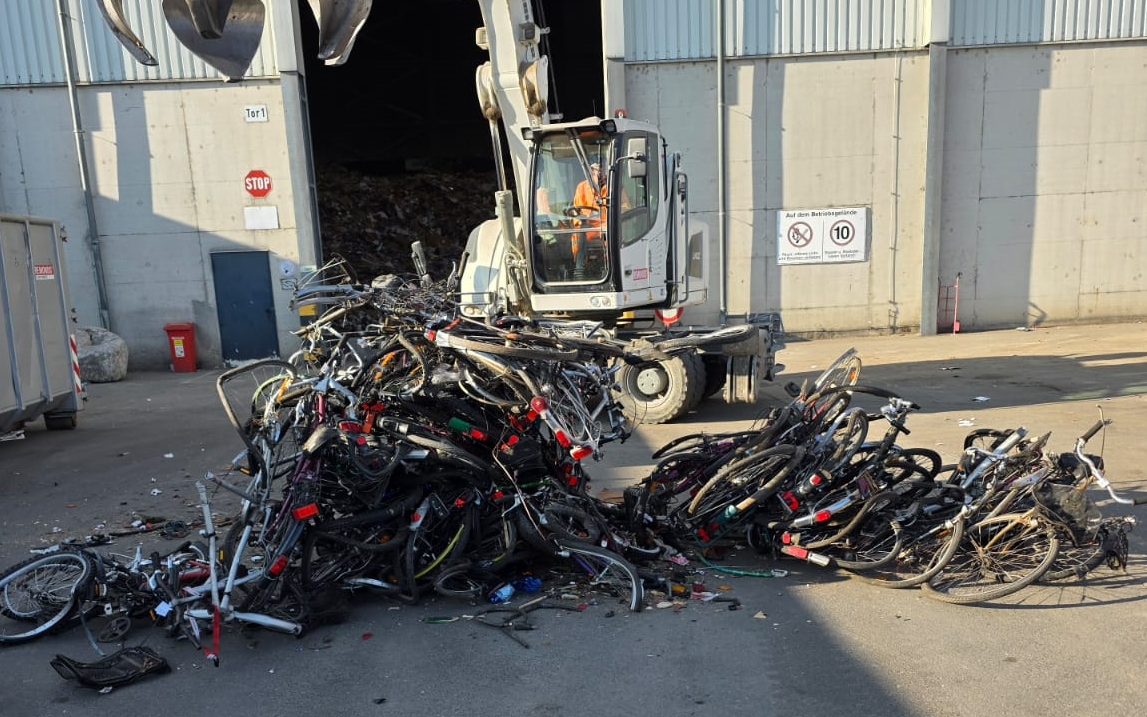 Ein Haufen Schrotträder liegen auf dem Boden. Im Hintergrund ist ein Bagger zu sehen.
