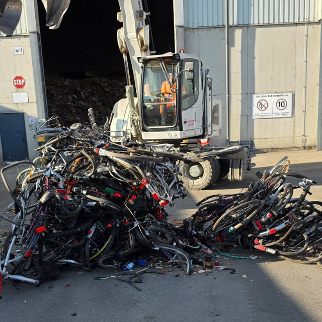 Ein Haufen Schrotträder liegen auf dem Boden. Im Hintergrund ist ein Bagger zu sehen.