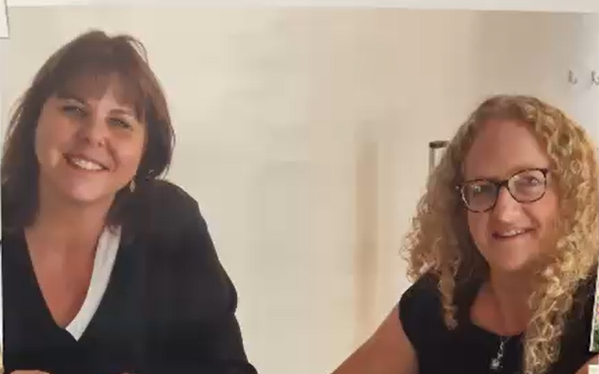 zwei Frauen sitzen an einem Tisch, stricken und schauen in die Kamera