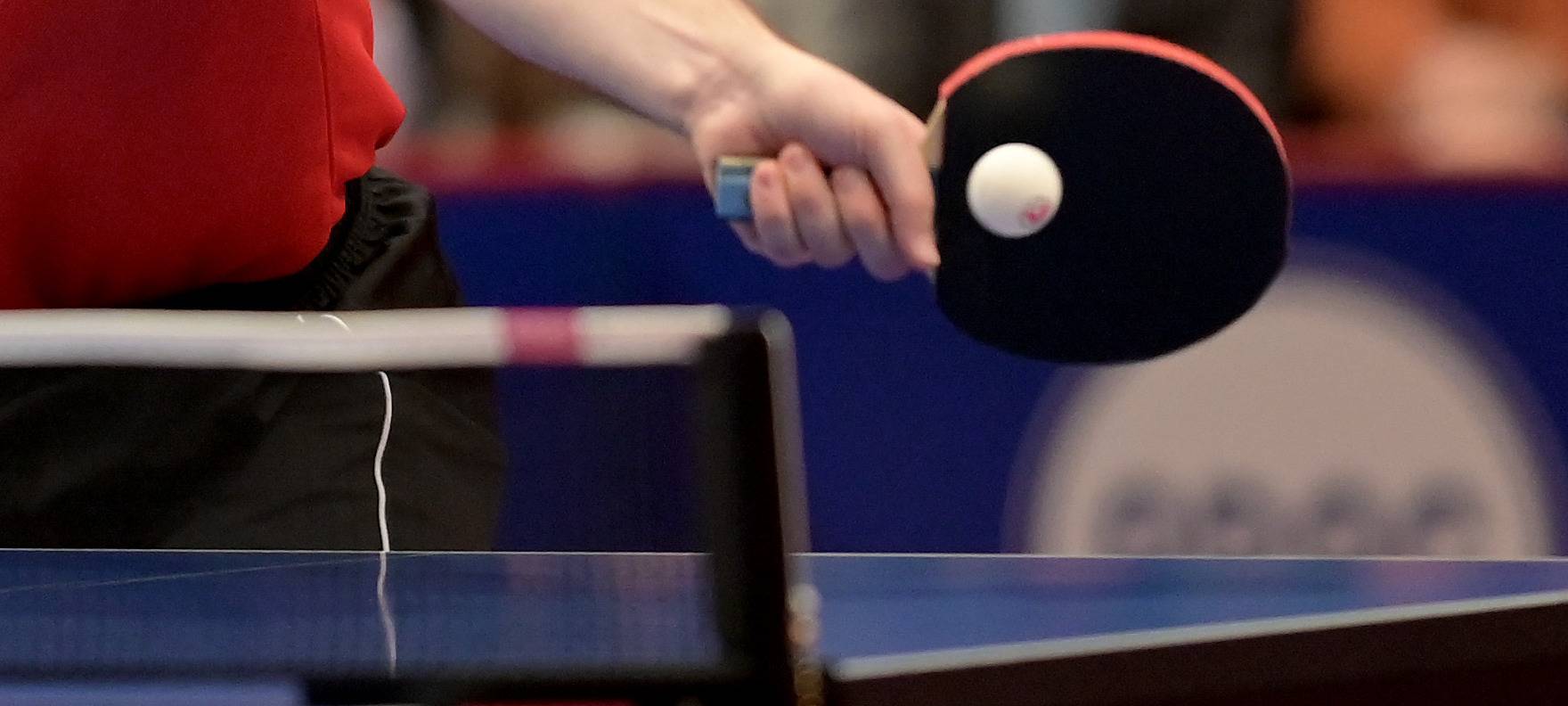 Ein Tischtennis-Spieler bei einem Tischtennis-Spiel. Im Fokus sind der Schläger, der Ball und das Netz.