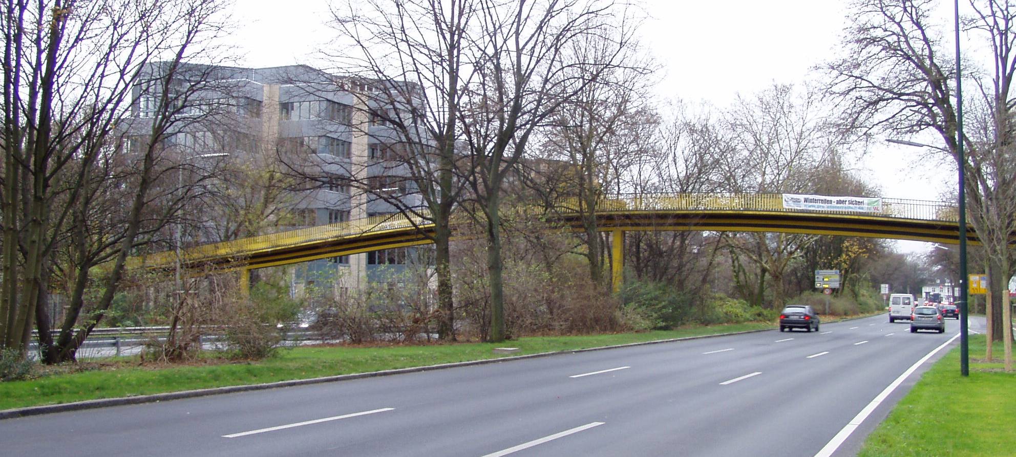 Blick auf den Kennedydamm in Richtung Norden. Über die Straße führt eine gelbe Fußgängerbrücke.