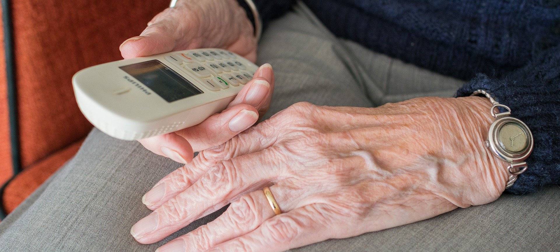 Die Hände einer älteren Person liegen im Schoß und halten ein Telefon
