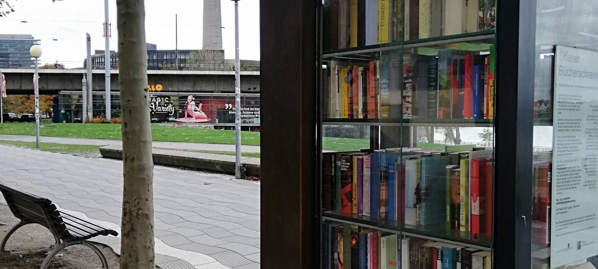 Ein Bücherschrank mit gebrauchten Büchern am Rheinufer. Im Hintergrund ist der Rheinturm zu sehen.