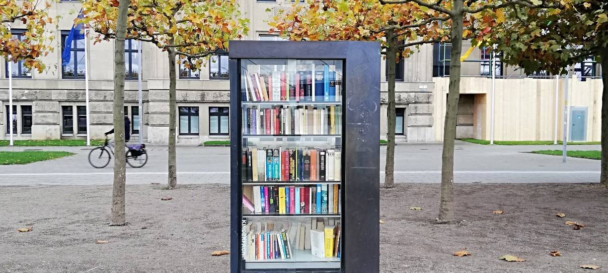 Ein Bücherschrank mit gebrauchten Büchern am Rheinufer. Im Hintergrund sind Bäume zu sehen.