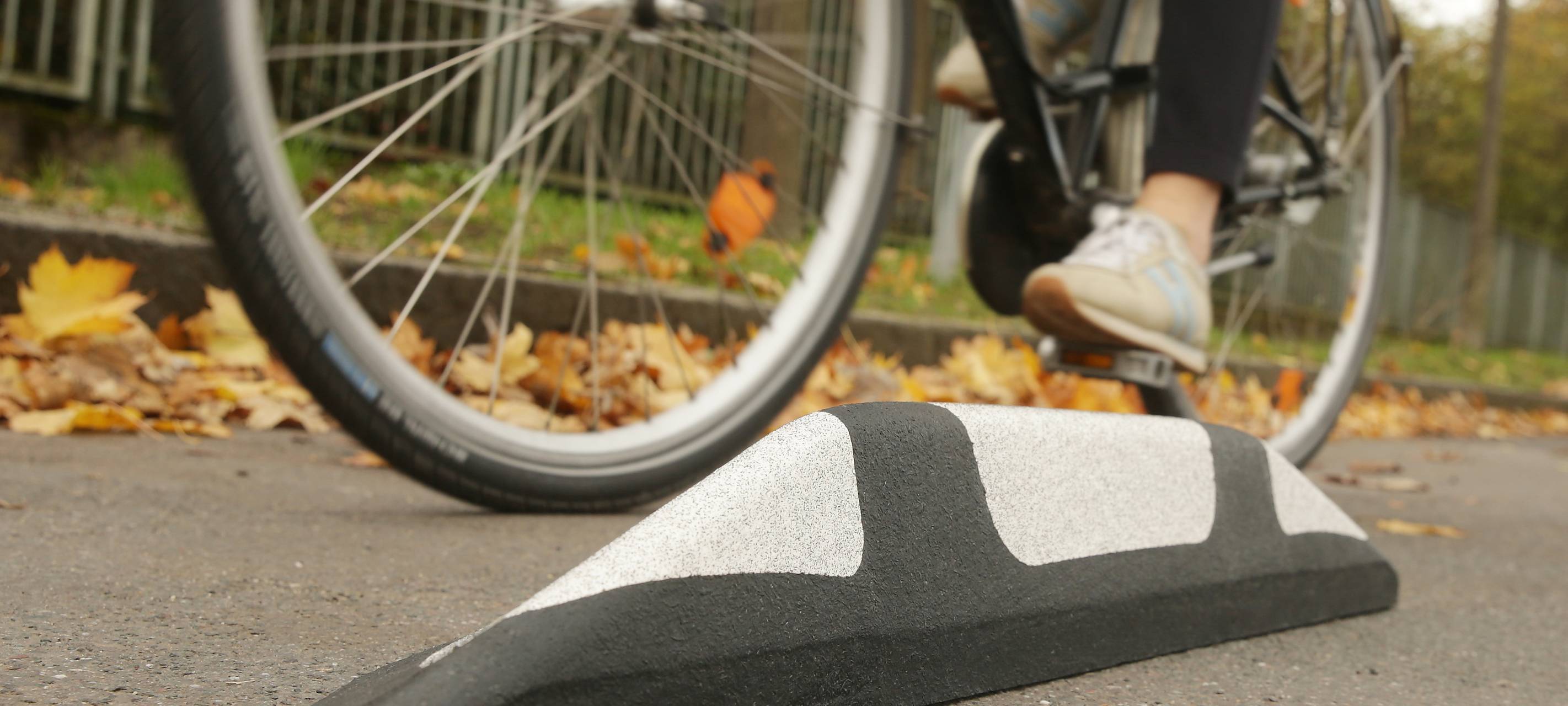 Zu sehen ist aus der Froschperspektive eine Person, die auf einem Fahrrad vorbeifährt. Am Straßenrand liegt Herbstlaub.