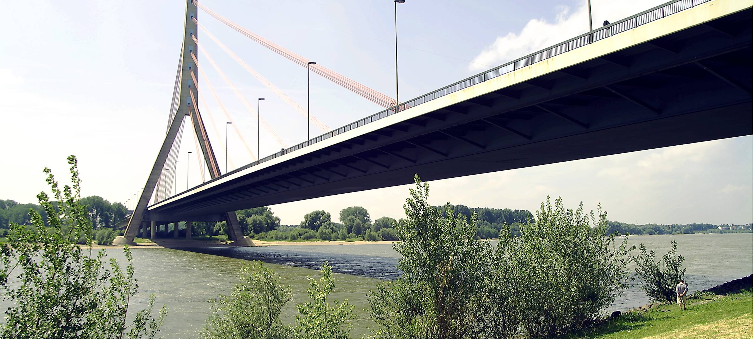 Die Fleher Brücke von unten fotografiert. Davor sind Teile vom Deich und dem Rhein zu sehen. Am Rhein sind grüne Sträucher.