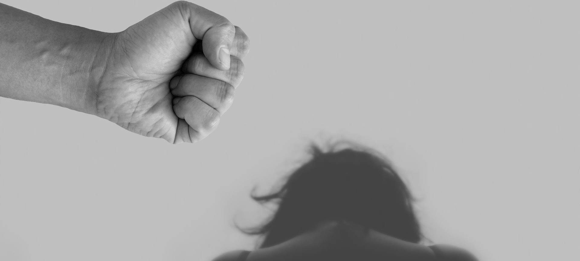 Eine Fraue ist zusammengesunken zu sehen. Eine männliche Faust ist drohend zu sehen.