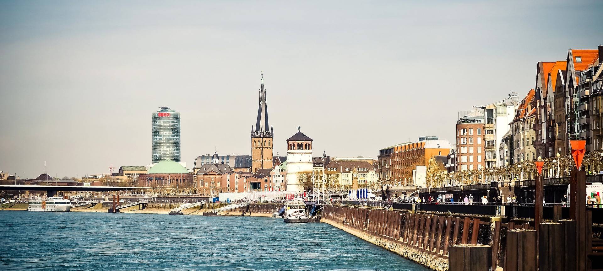 Ausblick auf die Düsseldorfer Rheinuferpromenade mit dem Schlossturm und der Basilika St. Lambertus im Hintergrund. Im linken Vordergrund ist der Rhein zu sehen.