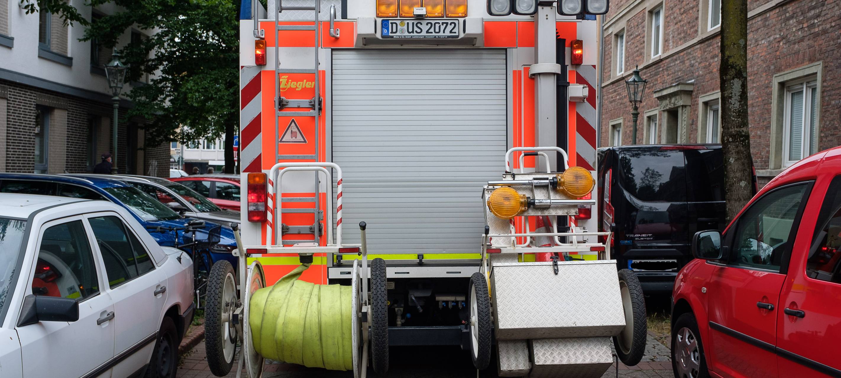 Die Rückseite eines großen Feuerwehrwagens. Links und rechts stehen Wagen. Der Feuerwehrwagen hat Schwierigkeiten, durchzukommen.