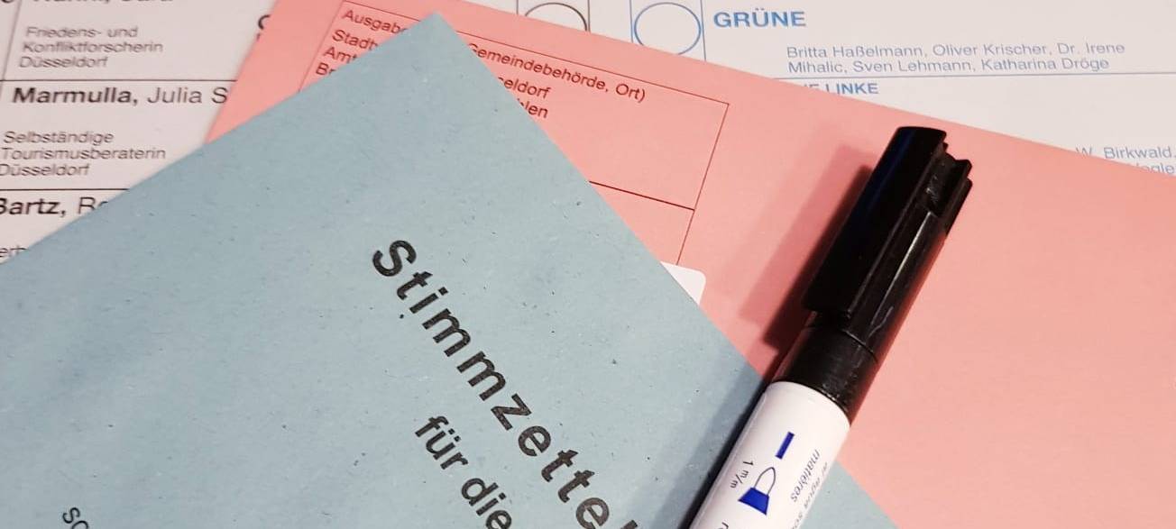 Die Briefwahlunterlagen - ein roter und ein blauer Umschlag liegen auf einem weißen Wahlzettel. Ganz oben liegt eins Stift zum wählen.