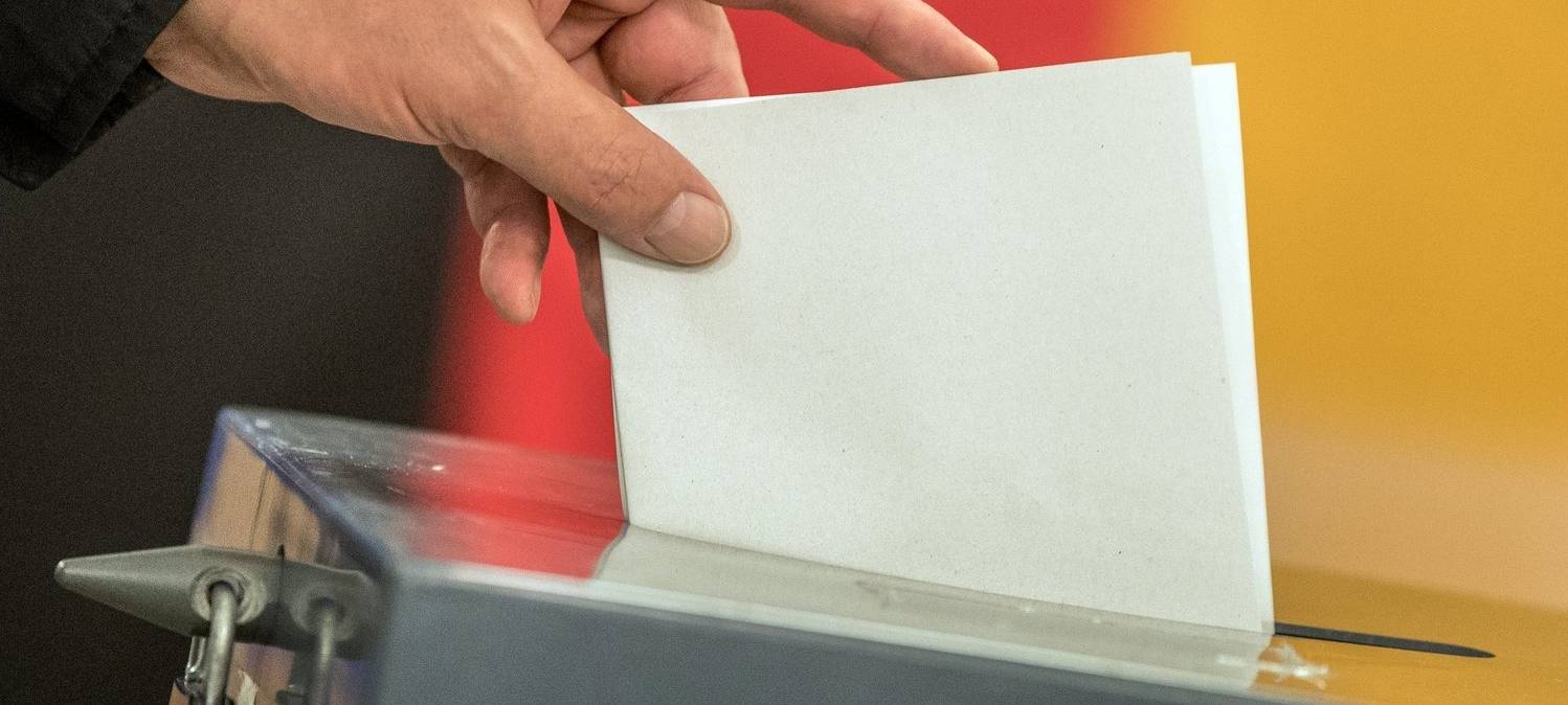 Ein Wahlzettel wird in eine Wahlurne eingeworfen. Im Hintergrund ist eine Deutschland-Fahne zu erkennen.