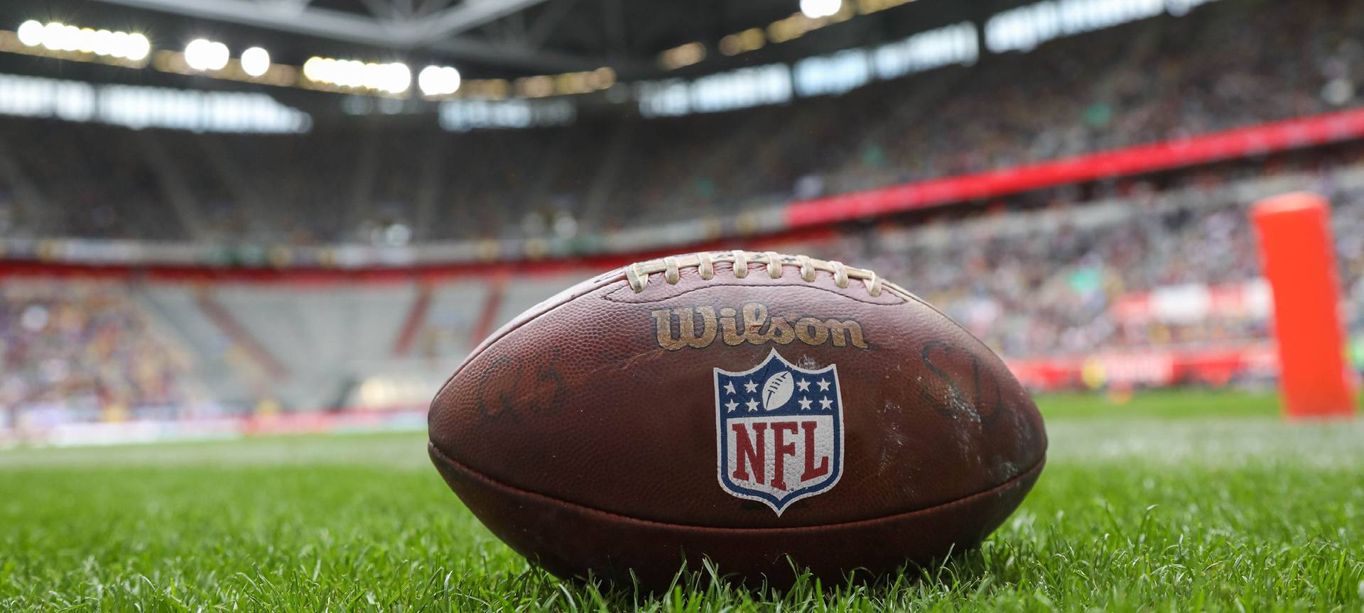 Im Vordergrund ist ein NFL-Football, er liegt auf der Wiese in einem Stadion.