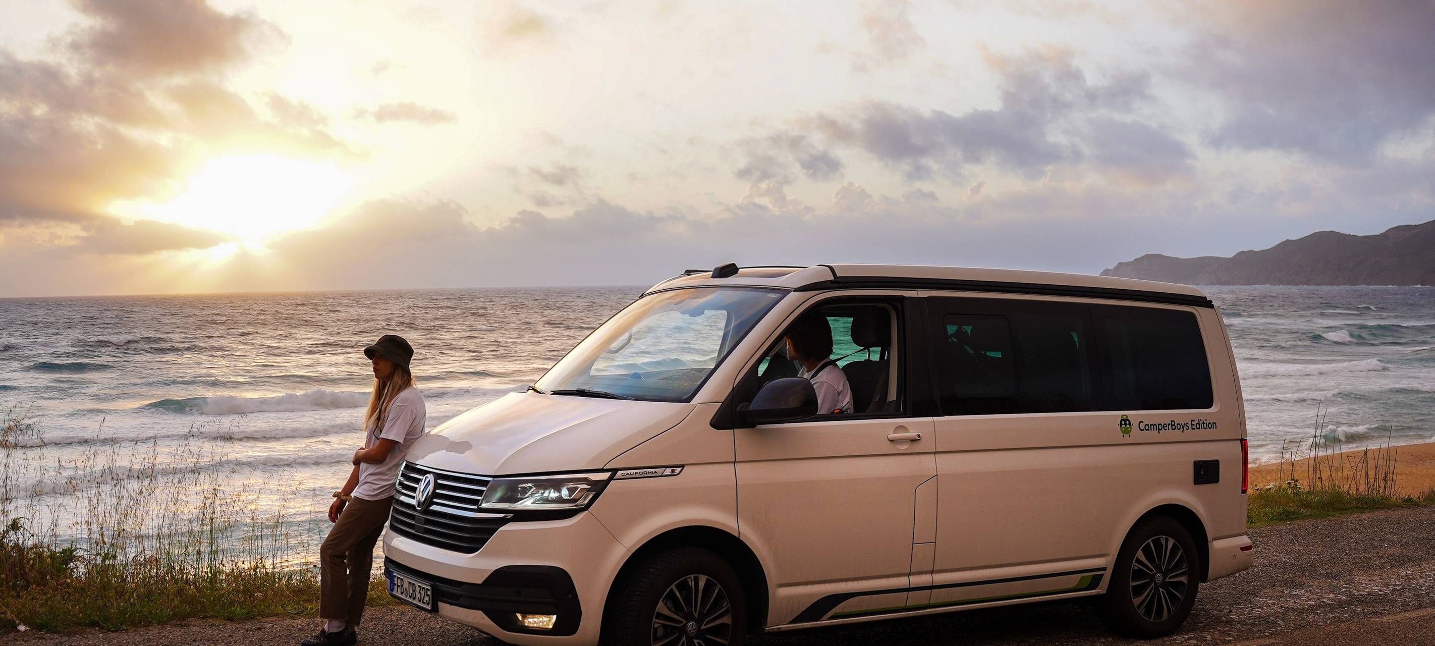 Ein Camper steht direkt am Meer. Die Sonne geht unter. Ein Mann lehnt entspannt am Wagen.