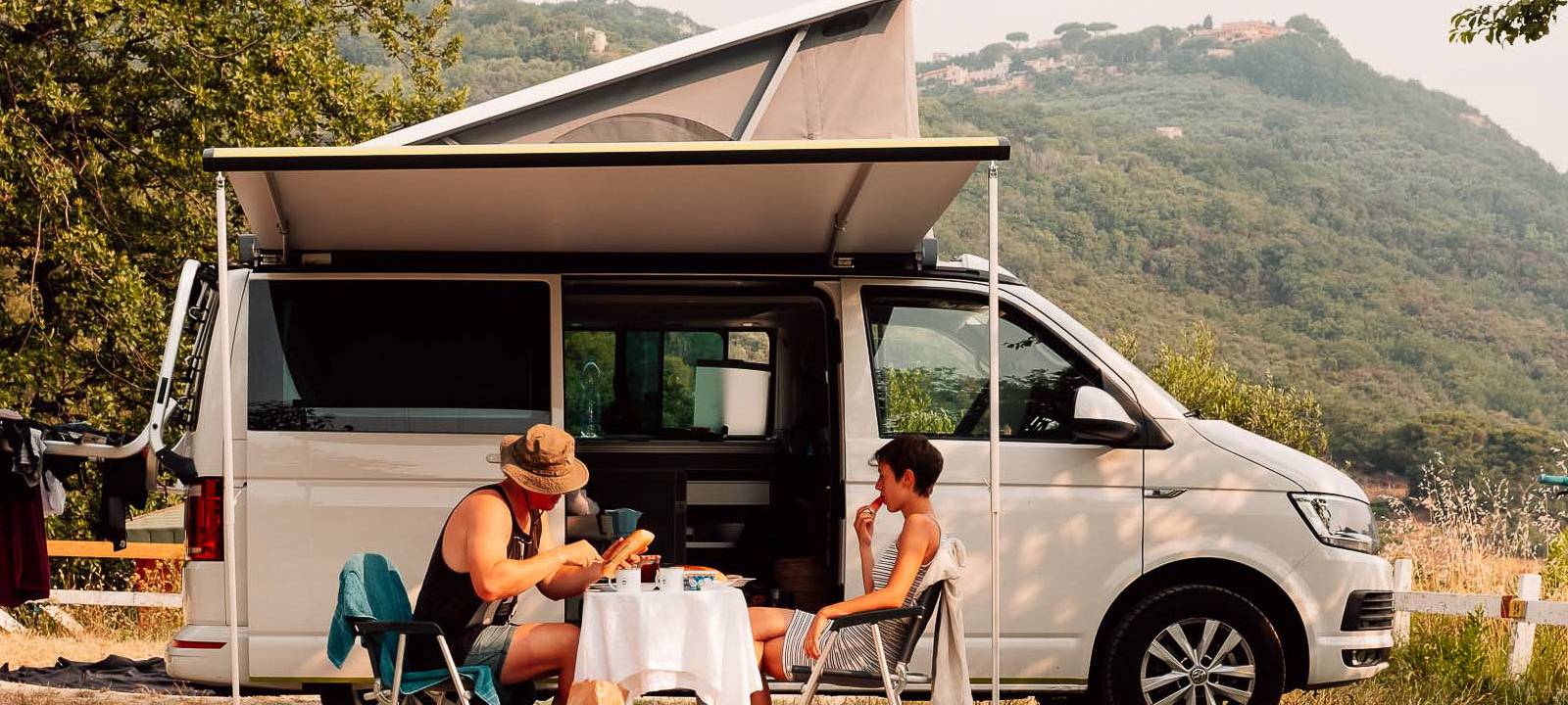 Eine Frau und ein Mann sitzen vor ihrem Camper in der Natur und essen.