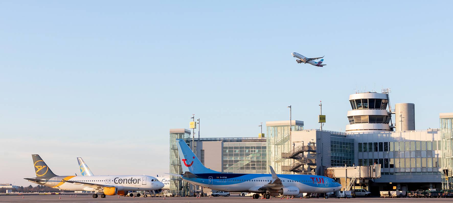 Eine Aufnahme vom Rollfeld des Düsseldorfer Flughafens. Zwei Flugzeuge stehen geparkt vor dem Flughafengebäude. Ein Flugzeug befindet sich in der Luft und startet gerade.
