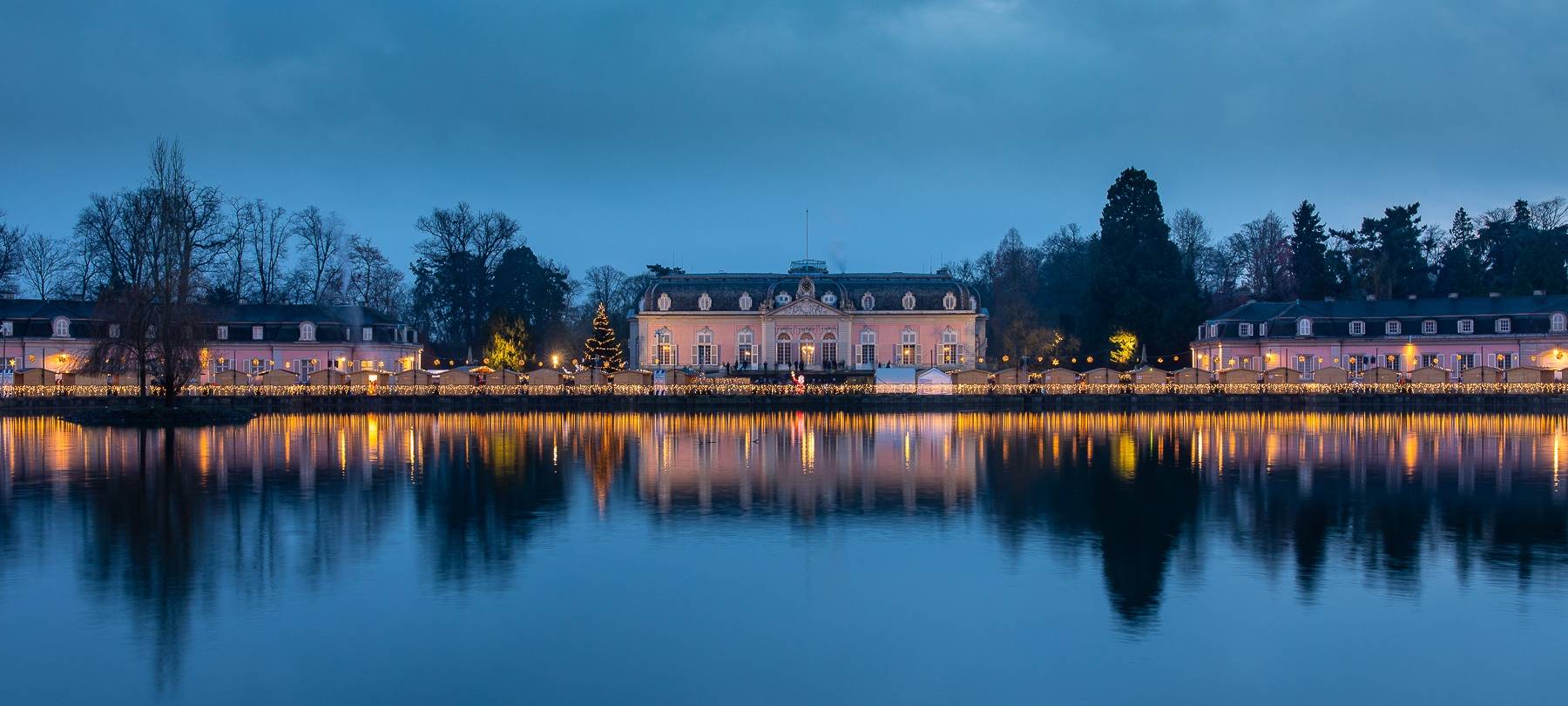 Stadtteil-Weihnachtsmärkte: Ein Weitblick auf den Weihnachtsmarkt vor dem Benrather Schloss. Es dämmert und die Buden sind festlich beleuchtet.