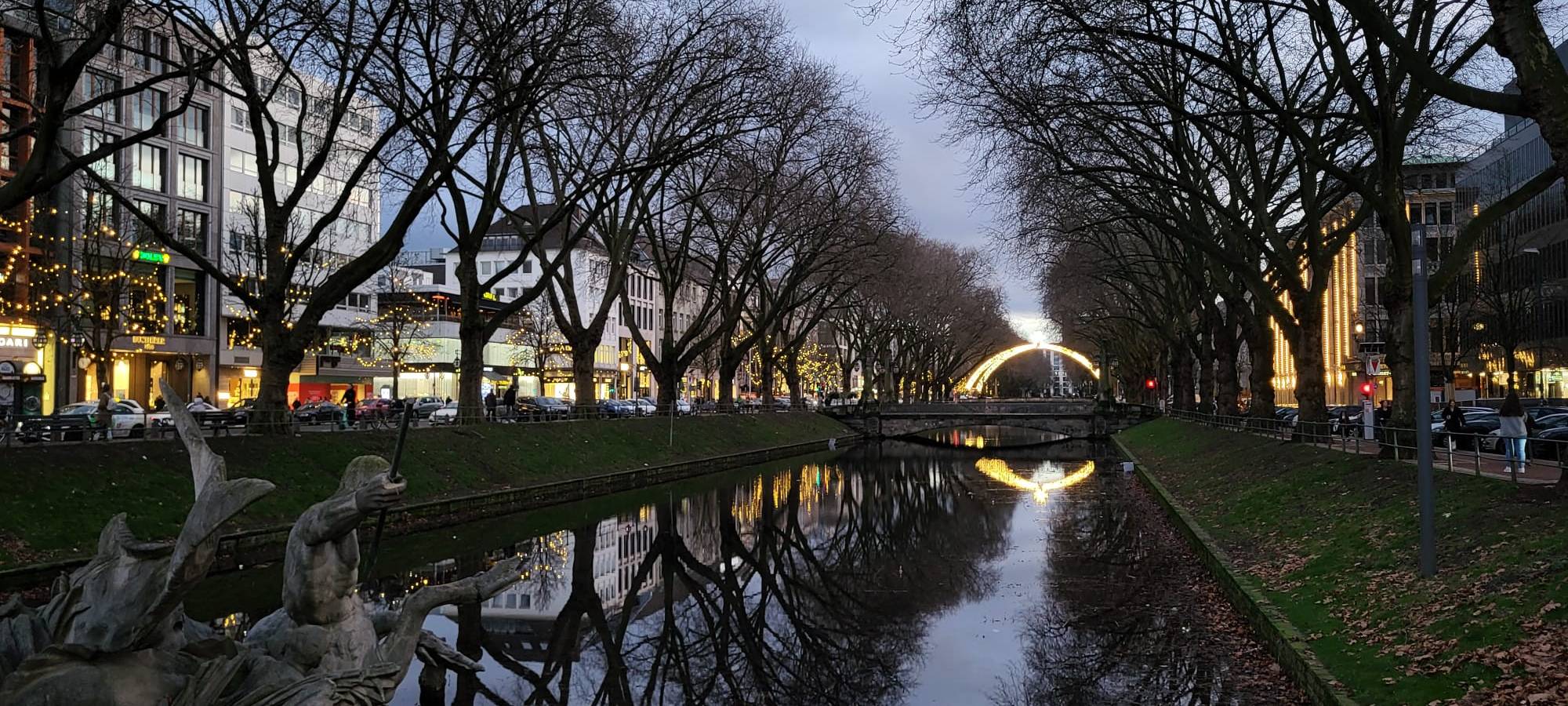 Die Königsallee mit dem Kö-Graben bei Nacht. Im Hintergrund leuchtet der Lichterdom auf der Steinstraße.