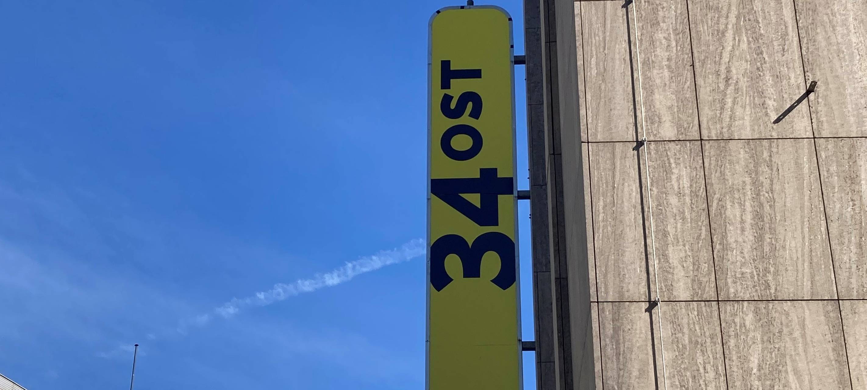 Ein gelbes Schild hängt draußen an einem grauen Gebäude. Darauf steht "34Ost".