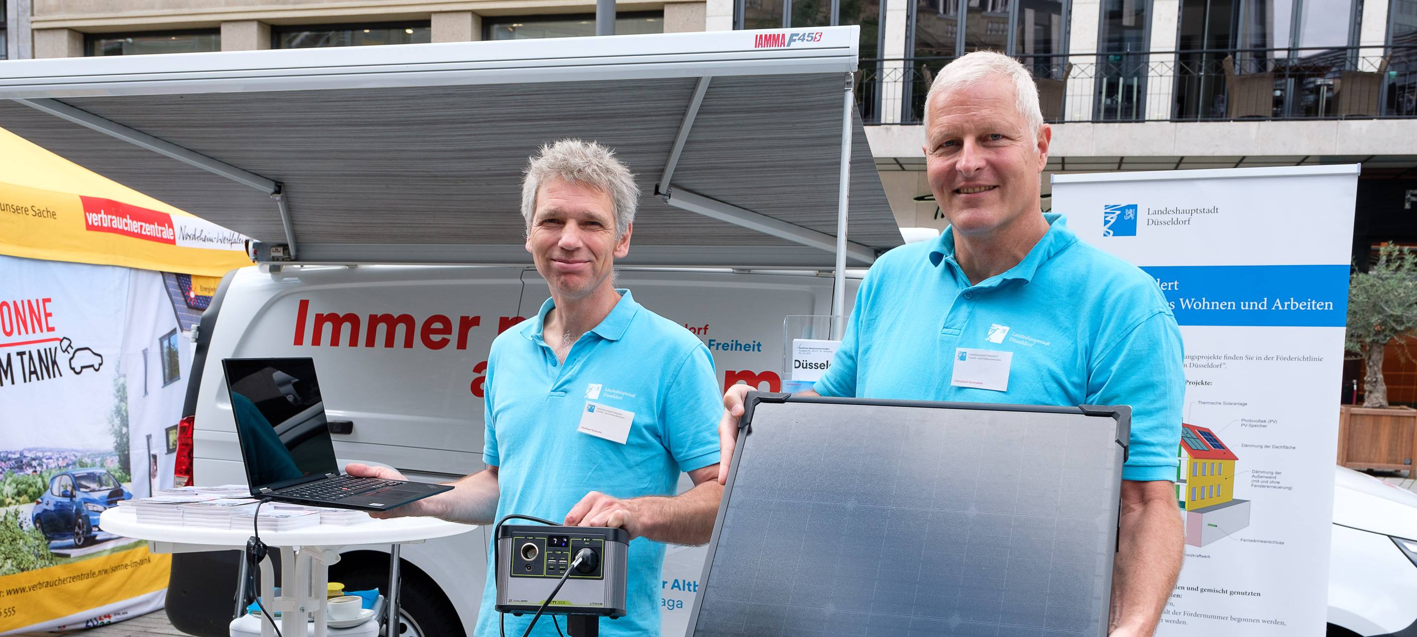 Zwei Männer im blauen T-Shirt stehen vor dem Klimamobil. Ein Mann hälte ein Solarpannel in der Hand.
