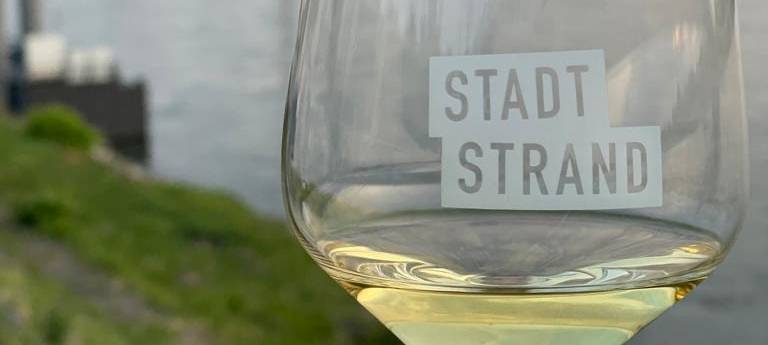 Ein Weinglas mit Weißwein und der Aufschrift "Stadtstrand", im Hintergrund ist die Düsseldorfer Skyline zu sehen