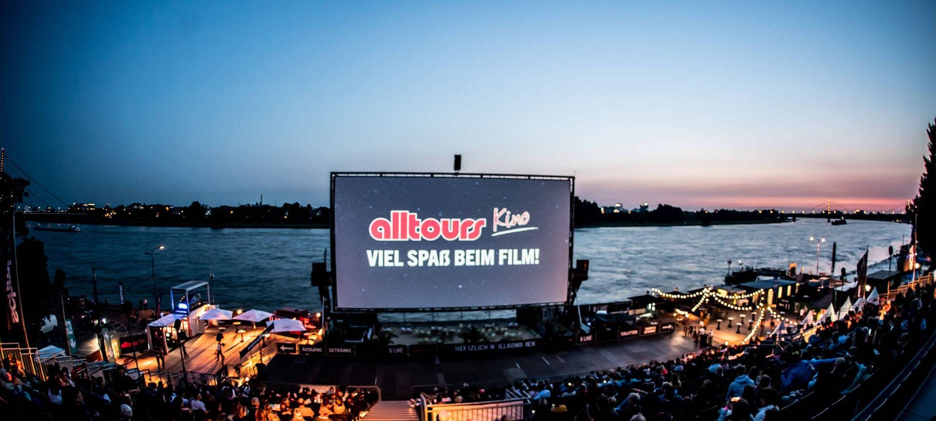 Zu sehen ist die Leinwand des alltours Open Air Kinos. Auf der Leinwand sieht man das alltours Logo. Im Vordergrund sieht man die Tribüne des Kinos, auf der viele Menschen sitzen - im Hintergrund sieht man den Rhein. Am Himmel geht grade die Sonne unter.