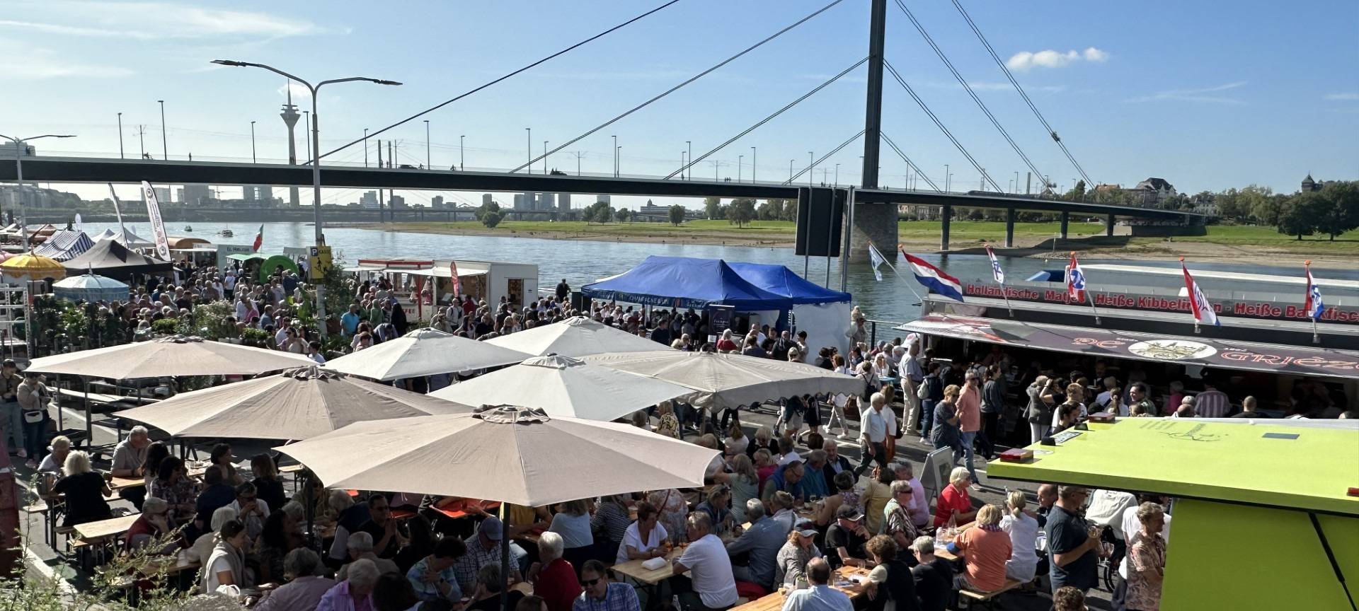 Blick auf den voll besuchten Fischmarkt am Düsseldorfer Rheinufer im Sonnenschein. Im Hintergrund ist die Oberkasseler Brücke und der Rhein zu sehen.