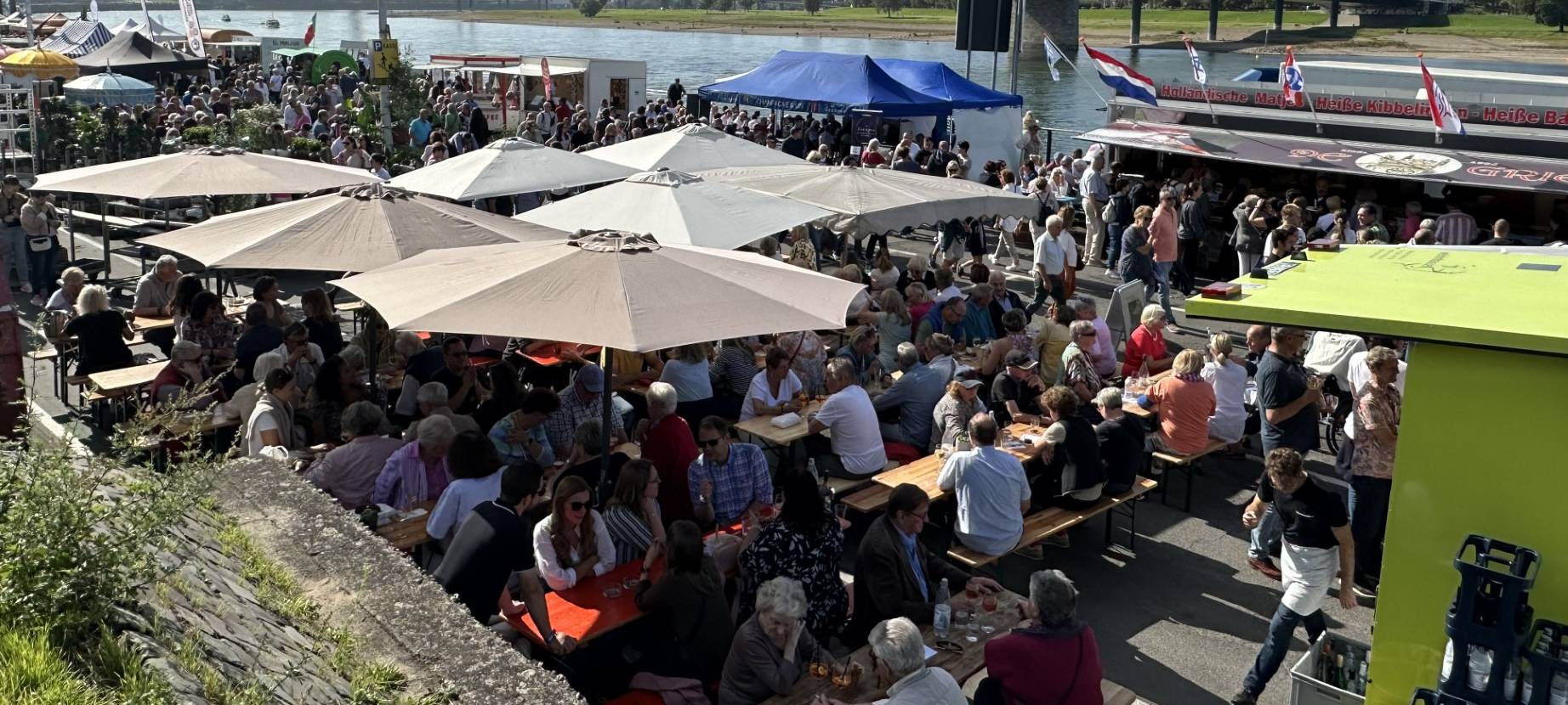 Blick auf den voll besuchten Fischmarkt am Düsseldorfer Rheinufer im Sonnenschein. Im Hintergrund ist die Oberkasseler Brücke und der Rhein zu sehen.