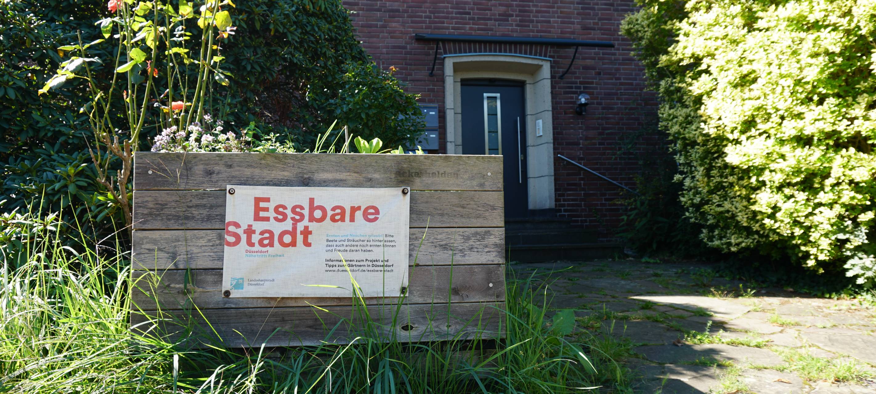 Blick auf ein Hochbeet der Stadt vor einem Wohnhhaus. Auf dem Hochbeet ist ein Papier mit dem Aufdruck "Essbare Stadt"