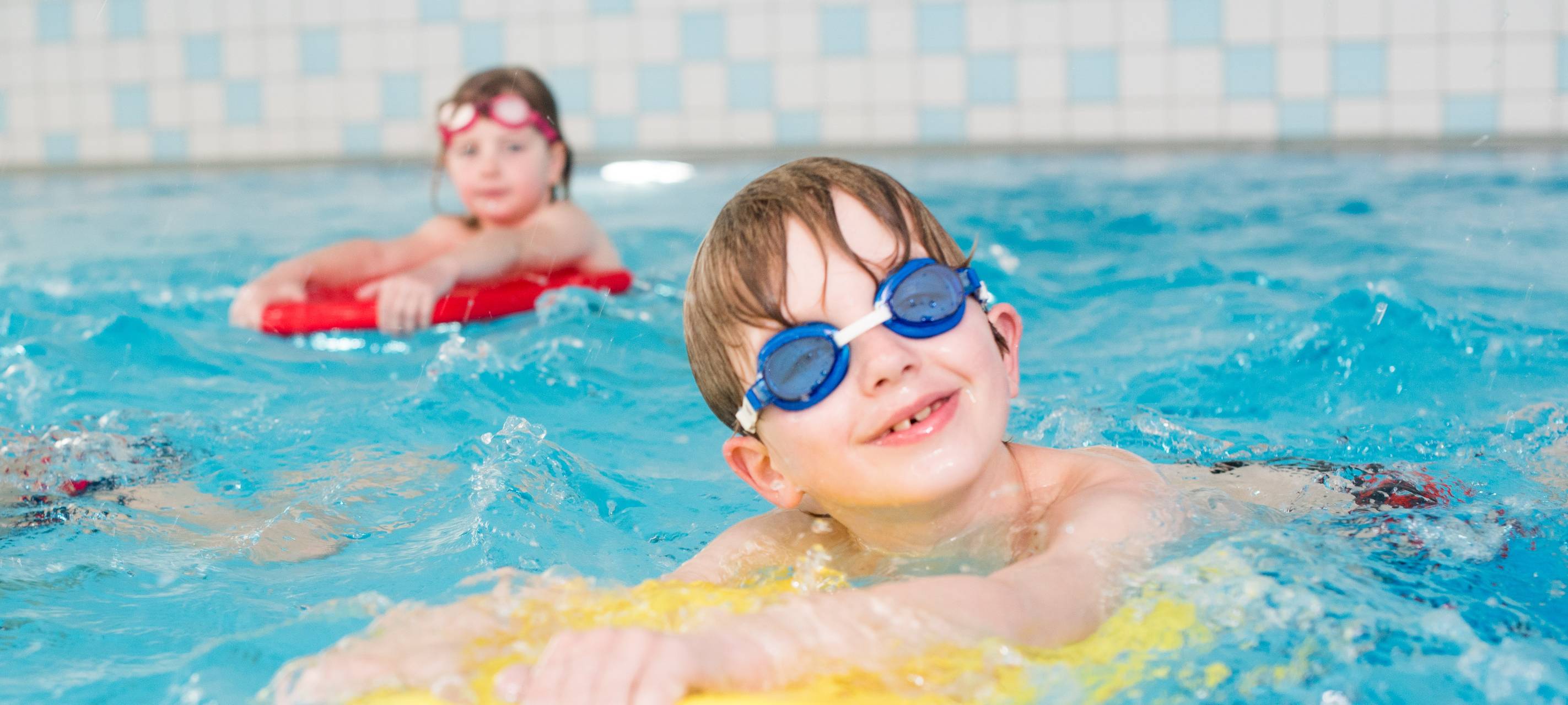 Ein Junge mit Schwimmbrille schaut in die Kamera. Er liegt auf einer gelben Schwimmhilfe.