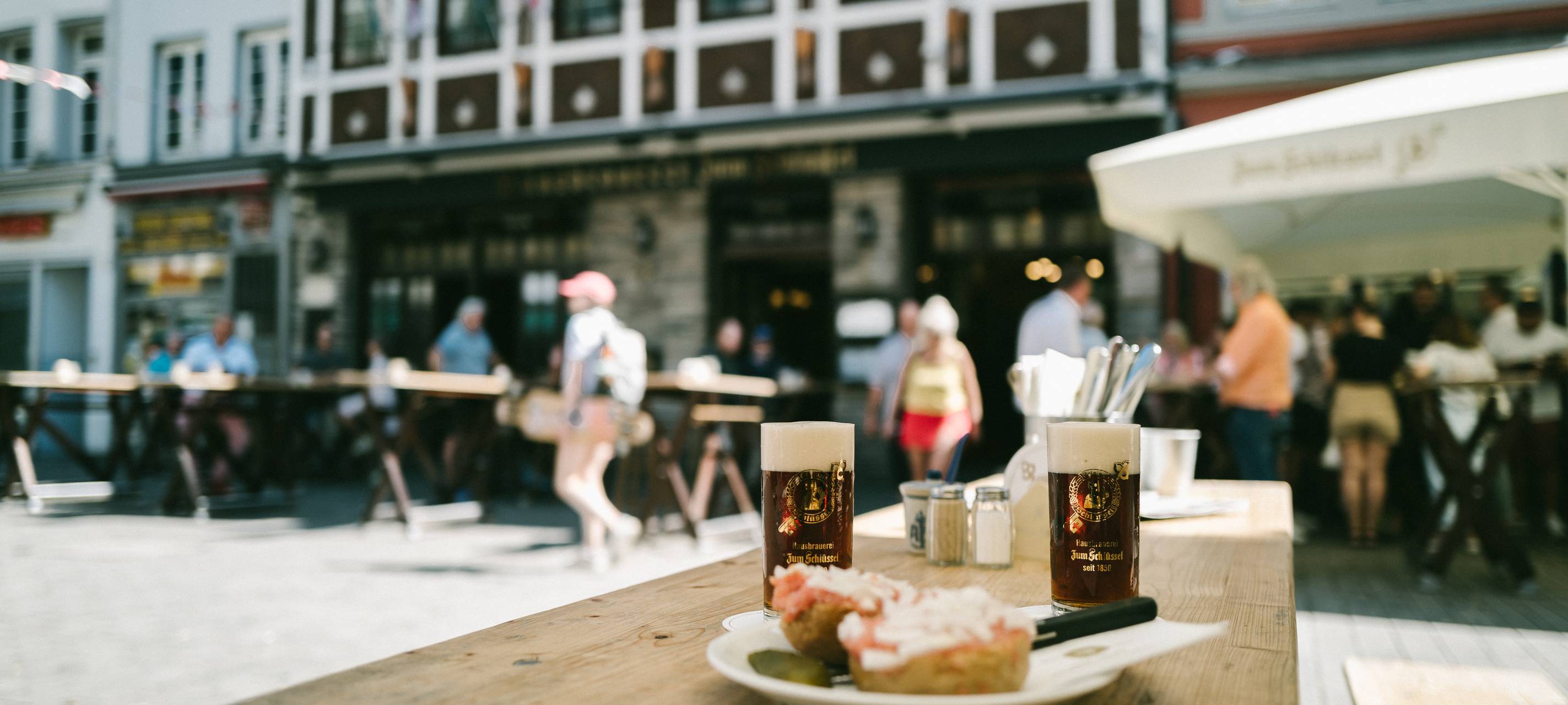Im Vordergrund sind zwei Gläser Schlüssel-Alt und ein Teller mit einem Mettbrötchen zu sehen. Im Hintergrund die Fassade der Brauerei Zum Schlüssel.
