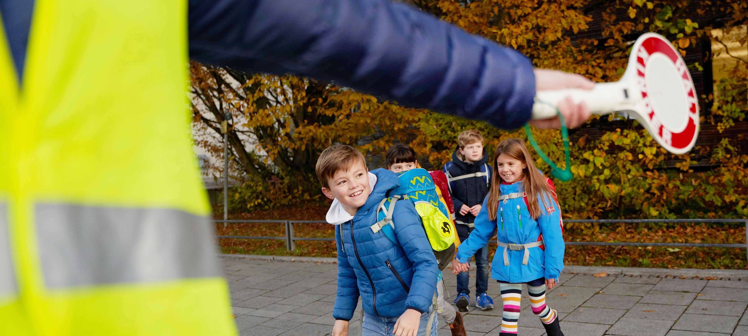I-Dötzchen auf ihrem Schulweg. Ein Ordner regelt mit einer Kelle den Verkehr und sorgt so für Sicherheit.