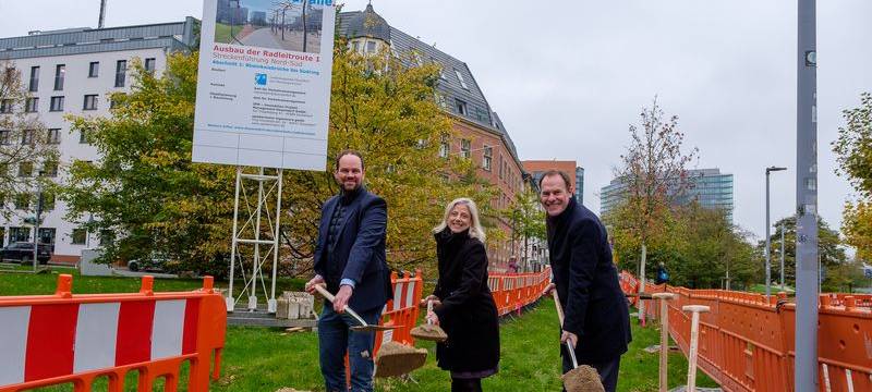 Der symbolische Spatenstich für die erste Radleitroute. OB Stephan Keller, Katharina Metzker Leiterin des Amtes für Verkehrsmanagement, und Michael Köhler, Geschäftsführer IPM Düsseldorf GmbH halten eine Schaufel in der Hand und stehen vor einem Sandberg.