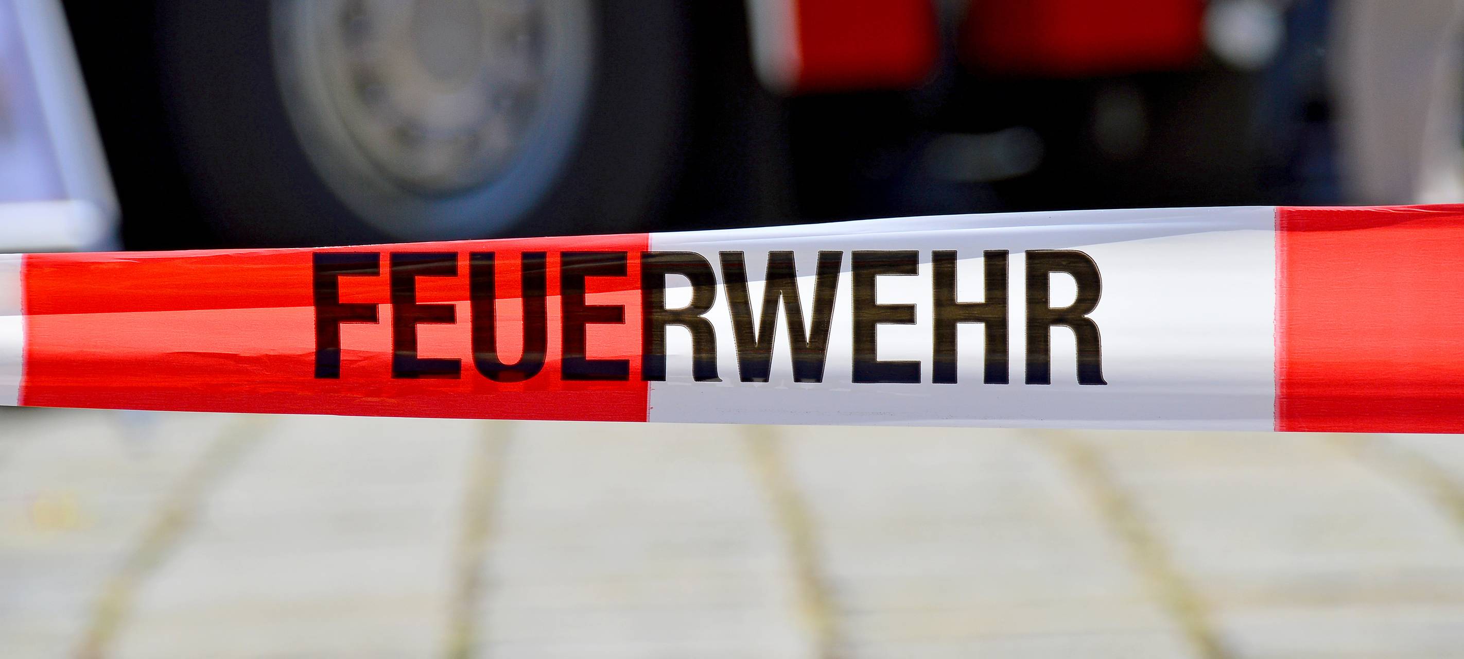 Ein rot-weißes Flatterband mit der Aufschrift "Feuerwehr". Im Hintergrund ist unscharf ein Feuerwehrwagen zu sehen.