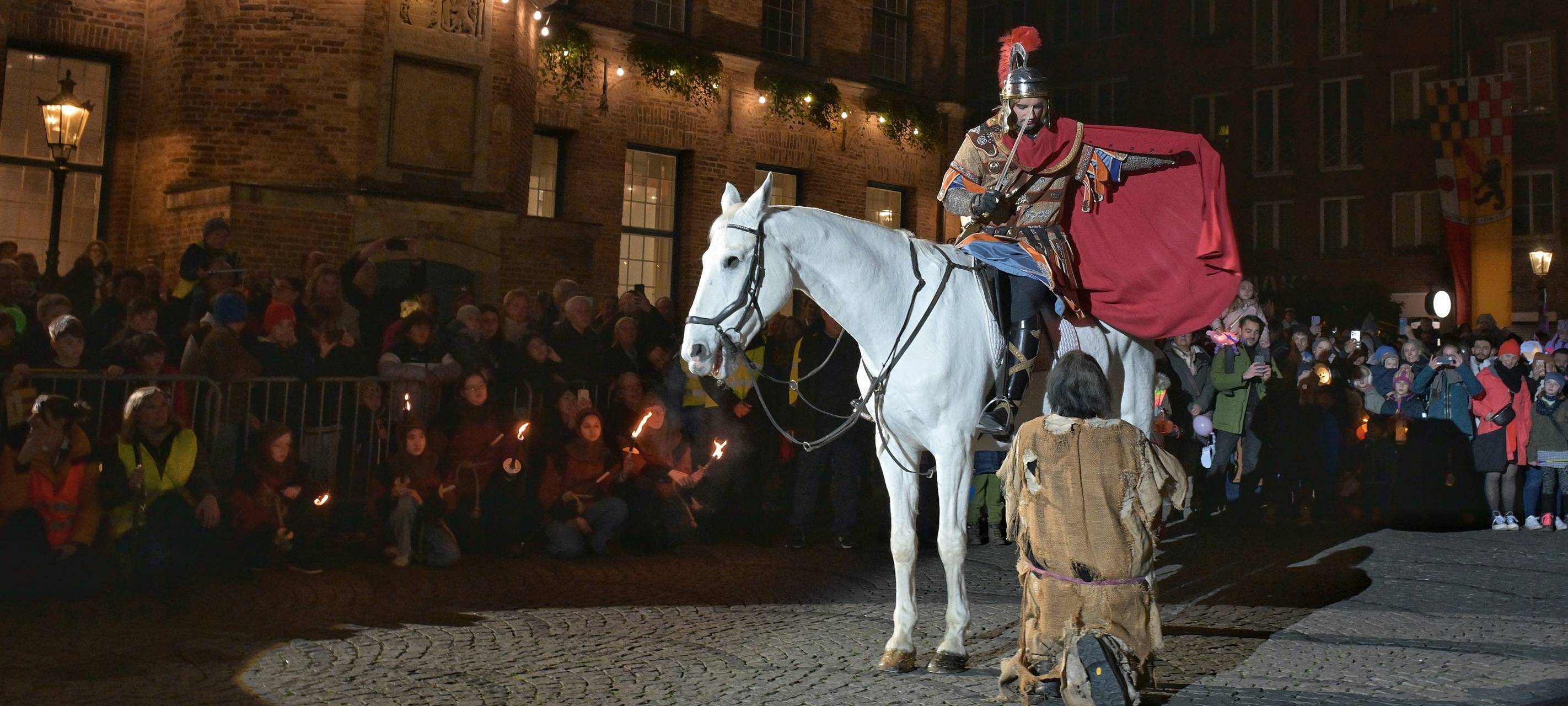 St. Martin teilt seinem Mantel mit einem armen Mann. Auf dem Martplatz wird das von vielen Menschen verfolgt.