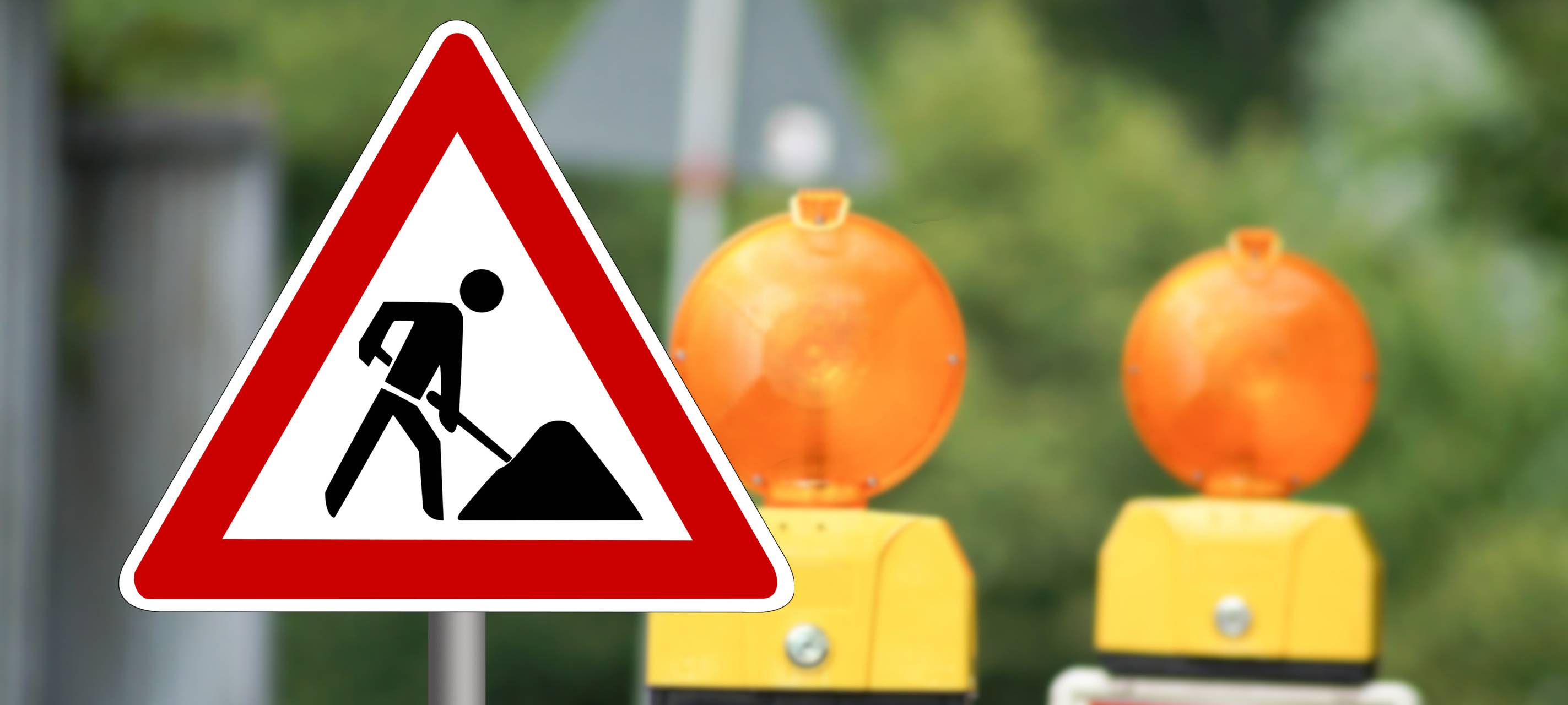 Ein Baustellenschild weist auf eine Baustelle hin. Im Hintergrund stehen zwei Warnbaken.