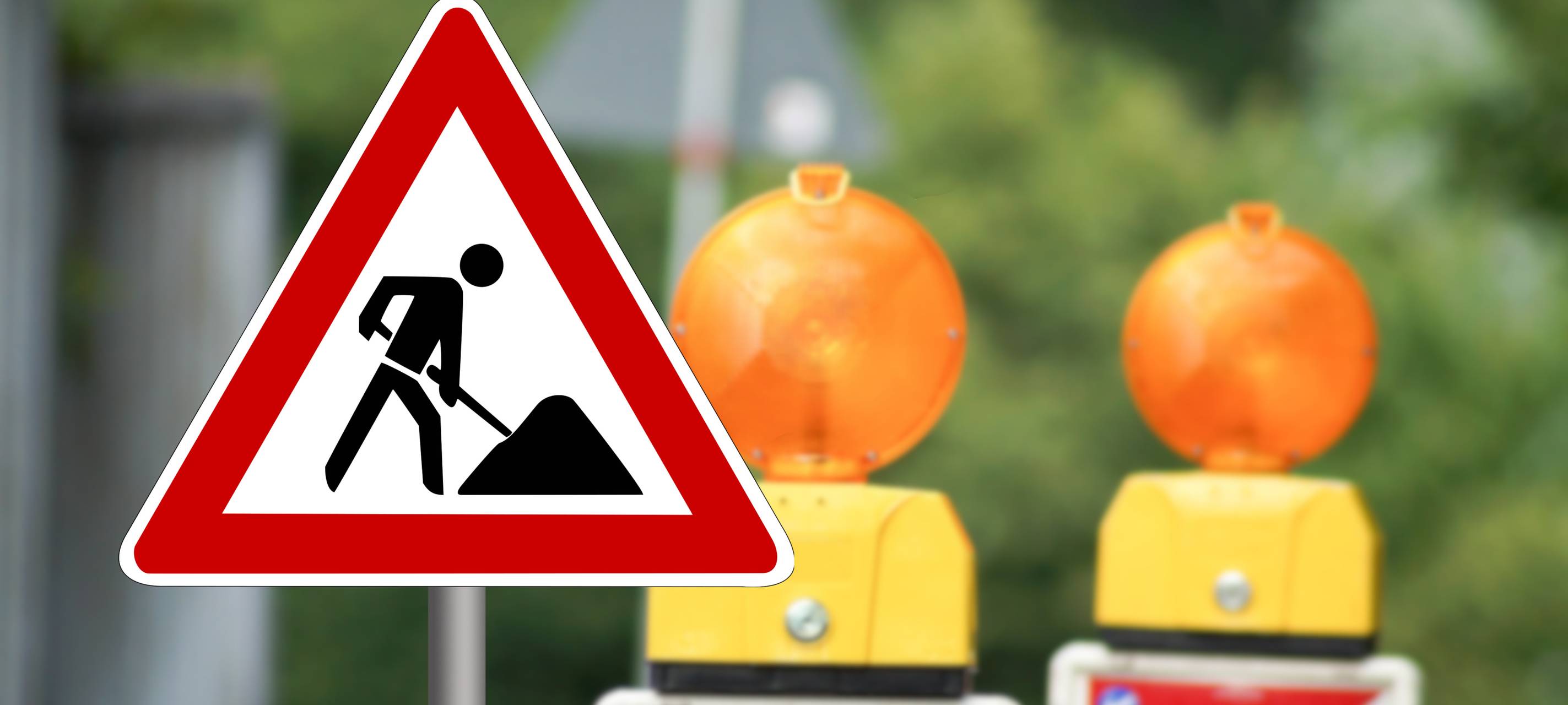 Ein Baustellenschild weist auf eine Baustelle hin. Im Hintergrund stehen zwei Warnbaken.