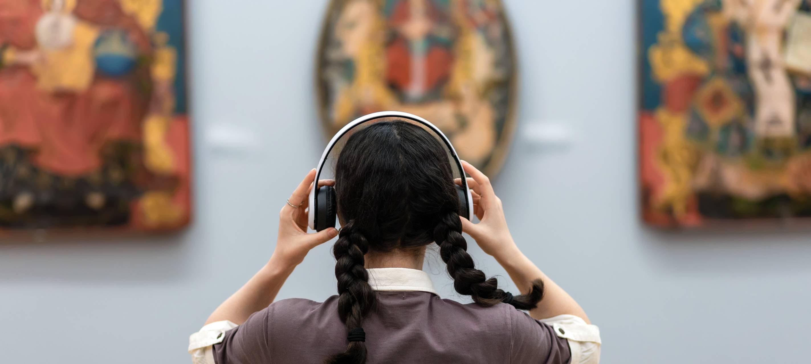 Eine Frau steht in einem Museum und schaut sich Gemälde an. Sie hat einen Kopfhörer auf; einen Audio-Guide.