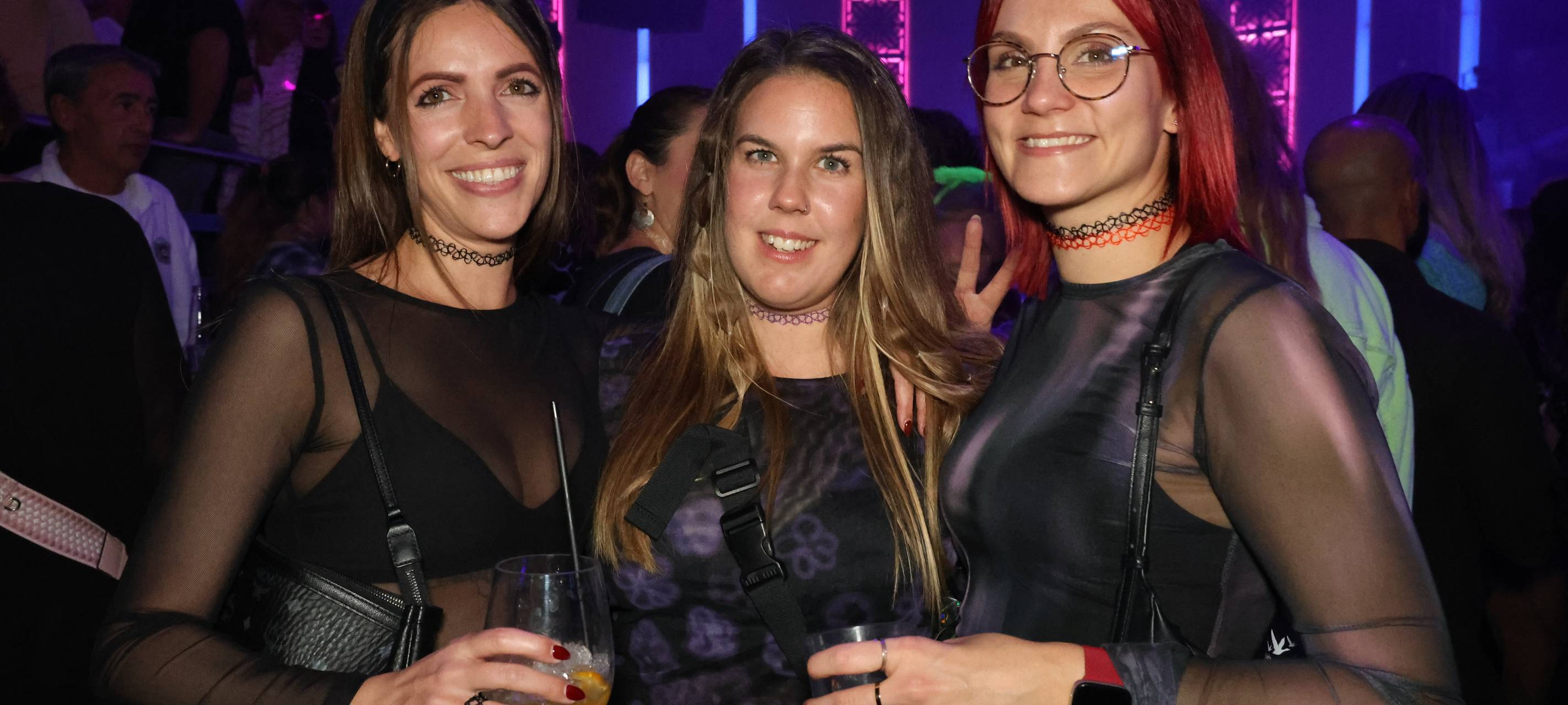 Drei Damen posieren für ein Foto in der Nachtresidenz. Sie tragen Tattoo-Halsbänder aus den Neunzigern und halten Getränke in der Hand.