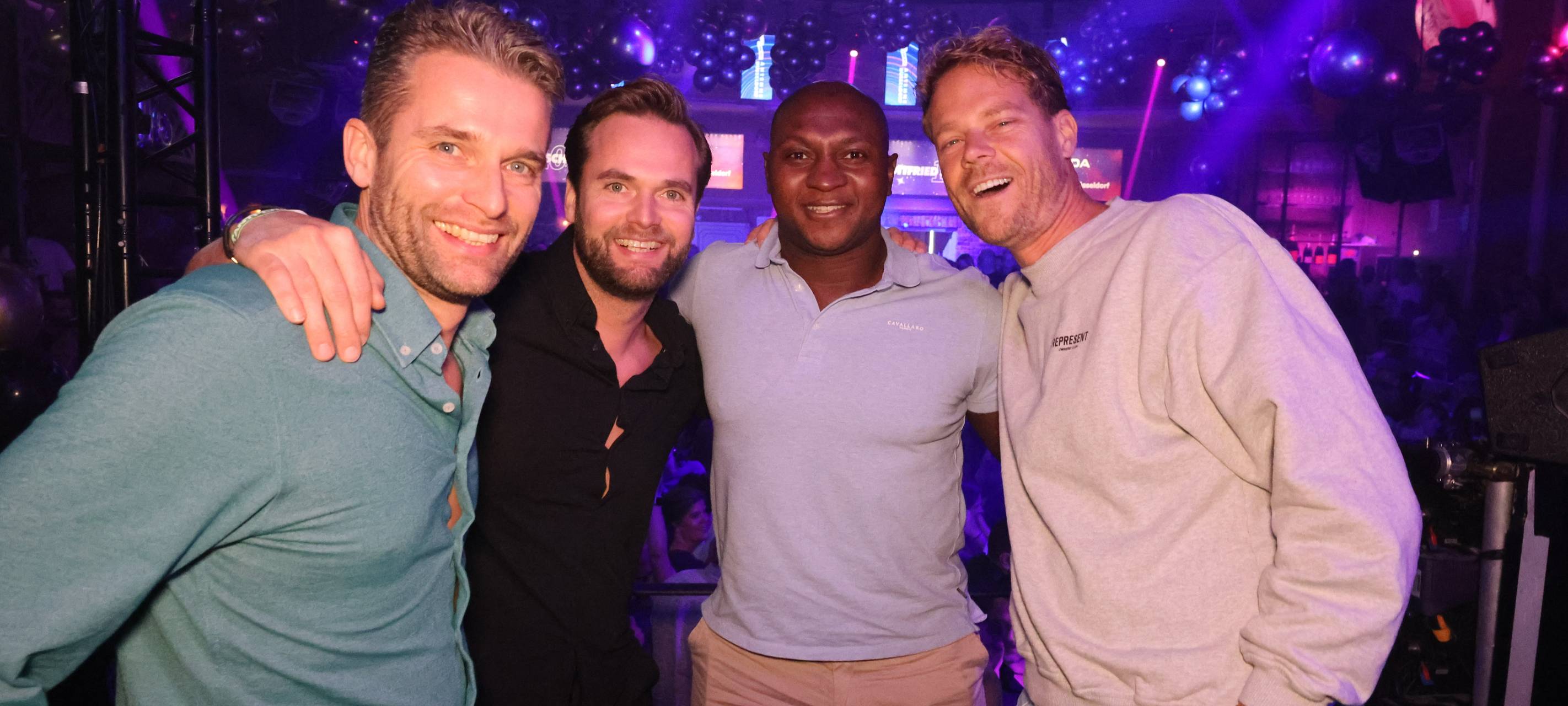 Vier Männer posieren gemeinsam für ein Foto in der Nachtresidenz. Im Hintergrund kann man Ausschnitte der Party sehen,