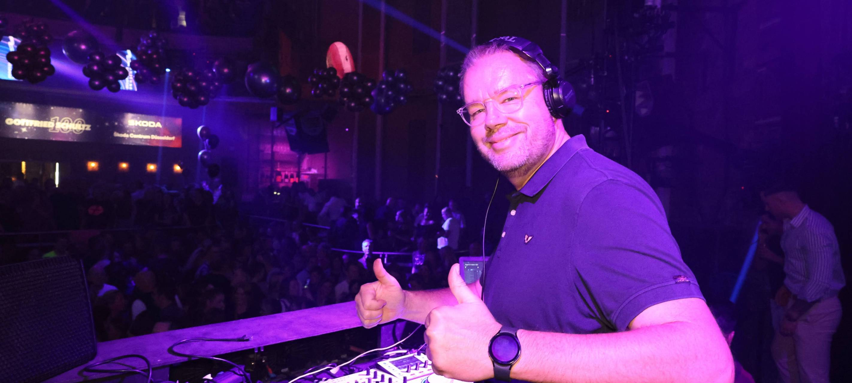 Antenne Düsseldorf-DJ Marc Pesch dreht sich zur Kamera und streckt seine Daumen nach oben. Er trägt Kopfhörer, neben ihm steht sein Mischpult. Im Hintergrund sieht man feiernde Menschen.