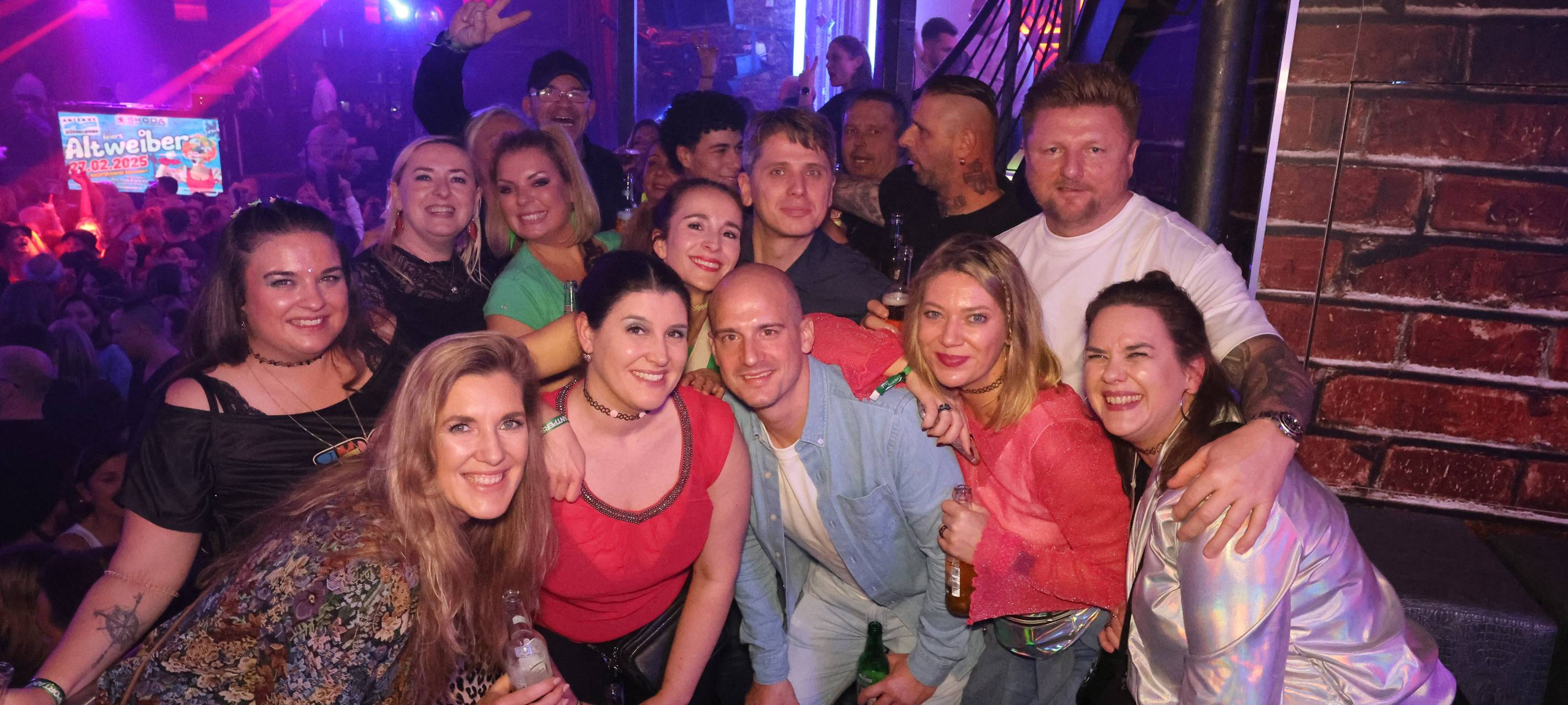 Eine Gruppe von siebzehn Personen (Männer und Frauen gemischt) posieren für ein Foto in der Nachtresidenz. Im Hintergrund sieht man feiernde Menschen.